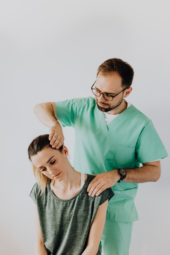 From above of bearded osteopath in eyewear and wristwatch stretching gently neck of young woman in casual clothes during medical examination in doctor office