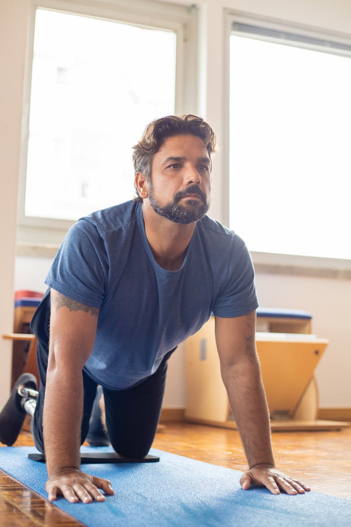 A determined man with a prosthetic leg kneels on a yoga mat for pilates in a bright room.
